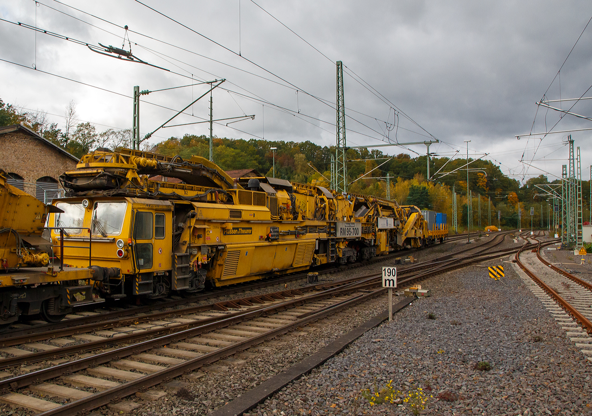 Die Plasser & Theurer Hochleistungs-Bettungsreinigungsmaschine RM 95-700, „Jacqueline“ , Schweres Nebenfahrzeug Nr. 99 80 9214 004-0 D-MGWB, ex 97 11 41 501 17-3, der MGW Gleis- und Weichenbau-Gesellschaft mbH & Co. KG (Berlin) am 22.10.2021 im Zugverband in Betzdorf (Sieg) bei einer Zugdurchfahrt.
 
Die Maschine wurde 2003 von Plasser & Theurer unter der Fabriknummer 4068 gebaut.

TECHNISCHE DATEN:
Bauart: RM 95-700
Spurweite: 1.435 mm (Normalspur)
Achsfolge:Bo´+Bo´Bo´+2´
Achsanzahl:  8
Länge: 48.240 mm
Drehzapfenabstände: 18.300 / 22.040 mm
Achsabstand im Drehgestell: 1.800 m 
Raddurchmesser: 920 mm (neu)
Höhe:  4.300 mm
Breite: 3.150 mm
Eigengewicht: 170,0 t
Motorengesamtleistung: 700 kW
V-Max Eigenfahrt:   20 Km/h
V-Max Zugverband: 100 Km/h
Zugkraft am Zughaken im Arbeitsbetrieb: ca. 300 kN
Streckenklasse:  D4
zur Mitfahrt zugel. Personen: 4
Anhängelasten (Eigenfahrt):  40 t 
Anhängelasten (Arbeitsfahrt bei 10 ‰ Steigung):  1.000 t
Bremsarten: KE G – P mZK und D2b 
Handbremsgewicht: 30 t 
Kleinster befahrbarer Radius: 120 m

Besondere Ausstattung:
• Laser-Anlage zur genauen Steuerung der Reinigungstiefe bei Ablage des Gleises in Hochlage
• Reinigungsbreite von 3.800 mm bis 5.000 mm durch Ergänzungsbalken
• maximale Veschiebung Gleisrost +/- 30 cm
• Leistung bis zu 700 m³/h
• Dokumentation der Lage des Gleises hinter der Reinigungsmaschine durch MKS-Messschrieb  
• Automatisches Warnsystem für Maschinenwarnung (AWS)
• Neuschotterzuführung zur optimalen Verfüllung des gereinigten Gleises mit Schotter

RM 95-700 - Bettungsreinigung mit Doppelsiebanlage
Die RM 95-700 ist eine achtachsige, vollhydraulische Hochleistungsbettungsreinigungsmaschine mit Neuschotterzuführung in Gelenkbauweise, für den Einsatz auf dem Gleisnetz der DB AG. Der Antriebs- und Siebwagen fährt vorne auf einem zweiachsigen und hinten auf vierachsigem Triebdrehgestell (2x2 Doppeldrehgestell). Der Aushub- und Einschotterteil ist vorne am Antriebswagen angelenkt und fährt hinten auf einem Laufdrehgestell.

Materialaushub
Die Aushubkette ist eine Weiterentwicklung der weltweit bewährten Ketten von Plasser&Theurer und wurde hinsichtlich der Lebensdauer weiter optimiert. Sie ist als endlose Fünfecks-Kratzerkette mit einer Höhe von 300 mm ausgeführt. Die Kettengeschwindigkeit ist stufenlos im Bereich von ca. 1,8 bis 3,5 m/sec verstellbar, die maximale Räumtiefe beträgt 1.000 mm unter SOK der überhöhten Schiene. Die Maschine ist mit einem Räumbalken für eine Reinigungsbreite von 4 Meter ausgestattet. Für geringere Räumbreiten wird ein Kettenquerbalken mit 3,4 m geliefert. Der hydraulische Kettenquerbalkenverschluss vereinfacht den Einbau des Querbalkens und verringert wesentlich die erforderlichen Rüstzeiten. Im Bereich der Räumkette ist die kontinuierlich arbeitende Gleishebe- und Rückeinrichtung angeordnet. Die maximale Hebung beträgt 250 mm, die maximale seitliche Gleisverschiebung ± 300 mm.

Siebanlage
Die Maschine ist mit zwei robusten Schwingsieben mit einer Gesamtfläche von über 40 m2 ausgestattet, die für höchste Qualität und Leistung sorgen. In der Überhöhung in Bögen verstellt der Maschinist die Querneigung der Siebanlage hydraulisch um bis zu 160 mm, sodass die Siebe immer waagrecht bleiben. Der Abraum gelangt über Förderbänder zur Maschinenfront. Das Übergabeförderband ist drehbar ausgeführt, sodass kein Schutzwaggon für Überstellfahrten notwendig ist. Der Abraum kann seitlich neben dem Gleis, auf am Parallelgleis stehende Waggons oder auf ein von der Maschine geschobenes Abraumverladesystem abgeladen werden.

Schotterverteileinrichtung
Der gereinigte Schotter gelangt von der Siebeinrichtung über Förderbänder in einen Schotterspeicher und von hier aus über ein Bodenförderband, einen Schotterschacht und ein Einschotterungsband direkt hinter der Räumkette in den Gleisbereich. Der Schotterspeicher und die Förderbänder auf der Aushubmaschine ermöglichen einen Mengenausgleich bei Arbeitsbeginn und Arbeitsende. Der Einschotterpflug mit verstellbaren Pflugschildern sorgt für eine gleichmäßige Verteilung des gereinigten Schotters.

Neuschotteranlage
Mit der RM 95-700 ist die Zuführung von Neuschotter im Zuge der Bettungsreinigung möglich. Dazu wird der Neuschotter von den hinten angereihten Materialförder- und Siloeinheiten in den Zuführungsschacht gefördert, und über die verstellbaren Einschotterhosen die Stopfzonen gezielt eingeschottert.

Lasereinrichtung, elektronischer Schreiber
Die präzise Führung der Räumkette in Längs- und Querrichtung ist von entscheidender Bedeutung für die Arbeitsqualität bei der Bettungsreinigung. Daher wird mit einer Lasereinrichtung die Bezugsbasis für die Steuerung von Räumtiefe und Kettenquerneigung vergrößert, um einen noch präziseren Schnitt zu gewährleisten. Diese Lasereinrichtung besteht aus einem Lasersender, der in einer Entfernung von bis zu 300 m (abhängig von Gleisgeometrie und Sichtverhältnissen) aufgestellt wird, und einem Laserempfänger an der Maschine. Der Empfänger wird entsprechend der Wahl des Bezugsstranges links oder rechts auf Konsolen an der Kettenführung positioniert. Die Voreinstellung der Räumtiefe erfolgt durch den Maschinisten von der Arbeitskabine aus. Hier steht auch eine Anzeige für Räumtiefe und ein Zeigerinstrument für die Planumsneigung zur Verfügung. Weiter ist die Maschine mit einem digitalen Vierkanal-Schreiber ausgestattet, womit folgende Parameter aufgezeichnet werden:
Räumtiefe
Planumsneigung
Überhöhung des gereinigten Gleises 
Verwindung des gereinigten Gleises

Kabinen
Alle Kabinen sind geschlossen, schallisoliert und vibrationsgedämpft ausgeführt. Gute Sichtverhältnisse sind gewährleistet. Kabinenheizung, Kabinenlüfter mit Luftfilter sowie Klimaanlagen (optional) sorgen für komfortable Bedingungen bei unterschiedlichen klimatischen Bedingungen während des Arbeitseinsatzes. In allen Kabinen ist außerdem eine Wechselsprechanlage installiert. Vorne und hinten befindet sich jeweils eine Fahrkabine mit den Bedienungselementen für Rangierfahrten. Direkt vor der Räumkette befindet sich die Arbeitskabine. Sie enthält sämtliche Bedienungselemente für die Kette, Hebe- und Rückeinrichtung, Arbeitsvorschub etc.

Die RM 95-700 ist eine Maschine für viele Einsatzmöglichkeiten
Mit der RM 95-700 steht eine Maschine zur Verfügung, die aufgrund ihres Konzeptes bei unterschiedlichen Baustellensituationen flexibel eingesetzt werden kann. Die RM 95-700 bietet dabei eine Reihe von Vorteilen:
• Hohe Arbeitsleistung durch die hohe Siebleistung
• Präziser Planumsschnitt durch Lasersteuerung
• Integrierte Neuschotterzuführung, damit ausreichender Schotter auch für die Ein- und Ausbaustelle und somit eine besser Nutzung der Sperrpausen
• Kurze Materialtransportwege innerhalb der Maschine
• Schotterspeicher für Materialmengenausgleich bei Baustellenanfang und Baustellenende
• Kurze Rüstzeiten durch hydraulischen Kettenquerbalkenverschluss und Montagekran
• Hohe Zugkraft durch drei angetriebene Drehgestelle
• Robuste Konstruktion des Maschinenrahmen
• Leistungsfähige Räumkette


Quelle: MGW Gleis- und Weichenbau-Gesellschaft mbH & Co. KG
