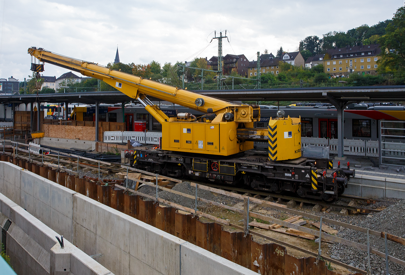 Nun im Einsatz der 125 t KIROW Gleisbauschienenkran KIROW Multi Tasker KRC 810 T (mit Schwerlastausrüstung), Schweres Nebenfahrzeug Nr. D-HGUI 99 80 9419 024-1, ex 97 82 53 513 19-3, der HERING Bahnbau GmbH (Burbach) ist am 29 September 2025 im Betzdorf/Sieg. 

Hier im Bereich unter dem P+R Parkhaus bis zum Fluss Sieg werden Horizontalbohrungen gemacht, vermutlich für erforderliche Entwässerungs-Leitungen. So mussten Bohrrohre in die Baugrube gehoben werden und wie hier der Abraum aus der Baugrube gehoben werden. Eigentlich ist ein 125 t Kran damit unterfordert (überdimensioniert), aber man muss bedenken es wurde eine weite Auskragung (Ausladung vor Puffer) und das freistehend (Nichtabgestützt) benötigt. 

Der hier mit der Ausladung vor Puffer von über 15 m könnte der Kran freistehend 30 t heben, aber darf dann nicht schwenken. Wenn geschenkt werden muss liegt diese dann nur bei 1,5 t. Entscheidend für die Ausladung des Auslegers ist aber auch die hintere Ausladung des Gegengewichts. 

Der Kran wurde 2008 von KIROW in Leipzig unter der Fabriknummer 149000 gebaut und an die HERING Bahnbau GmbH (Burbach) geliefert.

Der Gleisbauschienenkran KRC 810 T ist ein Schienendrehkran des Herstellers Kirow Ardelt (bis 2008 Kirow) in Leipzig. Der Typ 810 wird für den Aus-und Einbau von schweren Betonweichen, Gleisjochen und Hilfsbrücken sowie für die Montage von Ingenieurbauwerken eingesetzt. Bei Havarien auf Bahnstrecken leistet er schnelle Hilfe zum Bergen schwerer Lasten. Das maximale Lastmoment des Krans liegt zwischen 500 und 900 tm. Die Krane werden auch als Eisenbahndrehkran „EDK“ bezeichnet.

Die maximale Tragfähigkeit des bis zu 128 Tonnen schweren Krans beträgt abgestützt bis zu 125 Tonnen und freistehend 90 Tonnen. Bei maximaler Ausladung kann der Kran noch 28,5 Tonnen heben. Die größte mögliche Hakenhöhe liegt bei 18 m und die maximale Ausladung vor Puffer bei 15 m. Der Teleskoparm erlaubt Arbeiten im Radius zwischen 6,5 und 24,5 Metern. Im Gegensatz zu früheren Kranen ist auch ein Einsatz in der Nullposition ohne Abbau der Fahrleitung möglich. Die Gegenlast (Gegengewicht) ist zum Ausgleich unterschiedlicher Lasten ausfahrbar. Die Gegenlast kann in Transportstellung nicht (wie z.B. bei dem KRC 910) auf einem Gegenlastwagen abgestützt werden. Dadurch hat KRC 810 auch eine höhere Streckenlast (als z.B. der KRC 910). Für Hebearbeiten instehender Stellung verfügt der Kran über hydraulisch-ausfahrbare Stützen. Die Höchstgeschwindigkeit 19 km/h mit dem Eigenantrieb und 120 km/h im Zugverband geschleppt.  Zu dem Kran gehören zwei Kranschutzwagen. Die zulässige Anhängelast beträgt 80 t.

TECHNISCHE DATEN:
Spurweite: 1.435 mm (Normalspur)
Anzahl der Achsen: 8
Achsfolge: [(1A)(1A)][(A1)(A1)]
Länge über Puffer: 13.000 mm (ohne Ausleger)
Drehzapfenabstand: 8.000 mm
Drehzapfenabstand im Drehgestell 1 und 2: 2.300 mm
Achsabstand in den Einzeldrehgestellen: 1.100 mm
Ergebene Achsabstände in m: 1,1 / 1,2 / 1,1/ 4,6 / 1,1 / 1,2 / 1,1
Treib- und Laufraddurchmesser: 730 mm (neu) / 680 mm (abgenutzt)
Höhe in Transportstellung: 4.250 mm
Breite in Transportstellung: 3.100 mm
Eigengewicht: 128 t
Dieselmotor: Wassergekühlter 6-Zylinder Reihenmotor mit Abgasturboaufladung und Ladeluftkühlung vom Typ Cummins M 11 – C 315
Motorleistung: 235 kW bei 1.800 U/min
Max. Drehmoment: 1.458 Nm bei 1.300 U/min
Not-Dieselmotor: Luftgekühlter 2-Zylinder Viertakt-Dieselmotor mit Direkt-Einspritzung vom Typ Hatz 2 G 40 mit einer Leistung11,5 kW bei 2.300 U/min.
Fahrgeschwindigkeit im Zugverband: 120 km/h (geschleppt)
Fahrgeschwindigkeit mit Eigenantrieb: 20 km/h
zul. Anhängelast: 80 t
Hakenhöhe (max.): 18 m
Ausladung (max. vor Puffer): 16 m
Zugelassen als Schwerwagen für Streckenklasse: CS 05
Maximale Radsatzlast: 16 t
Streckenlast (Meterlast):  9,9 t/m
kleinster befahrbarer Radius: 00 m
Bremse: KE-GP-D- m.Z. (Scheibenbremsen auf allen Rädern)
Sekundär Scheibenbremsen auf allen Rädern
Handbremse: hydro-mechanisch auf alle Radsätze
Kraftstoffvorrat: 990 l

Tragfähigkeiten:
bei 0,00 m Ausladung vor Puffer, mit Schwerlastausrüstung: 125 t
bei 2,80 m Ausladung vor Puffer, ± 30°, abgestützt: 90 t
bei 2,80 m Ausladung vor Puffer, ± 20°, freistehend: 46 t